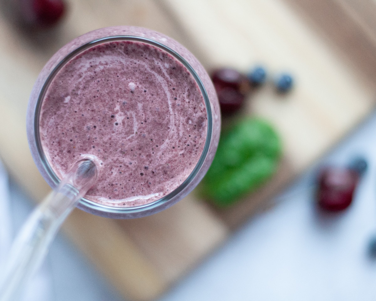 top down view of close up of a glass filled to the brim with this cherry blueberry smoothie with spinach. A glass straw is in the glass and ingredients are scattered around.