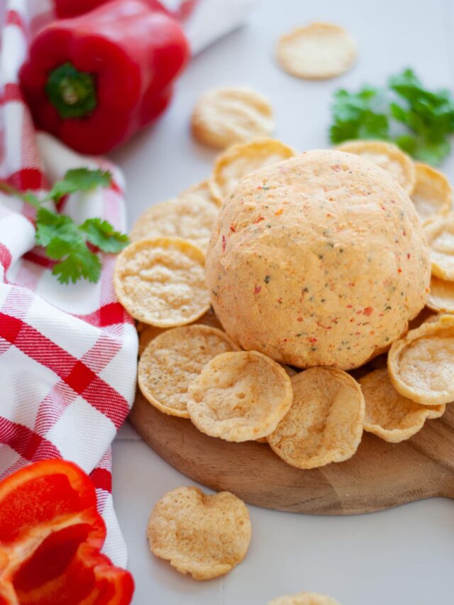 a tex mex cheese ball served on a round wooden platter with chips. there is cilantro, bell peppers, a red and white checked linen, and additional chips, around the scene.