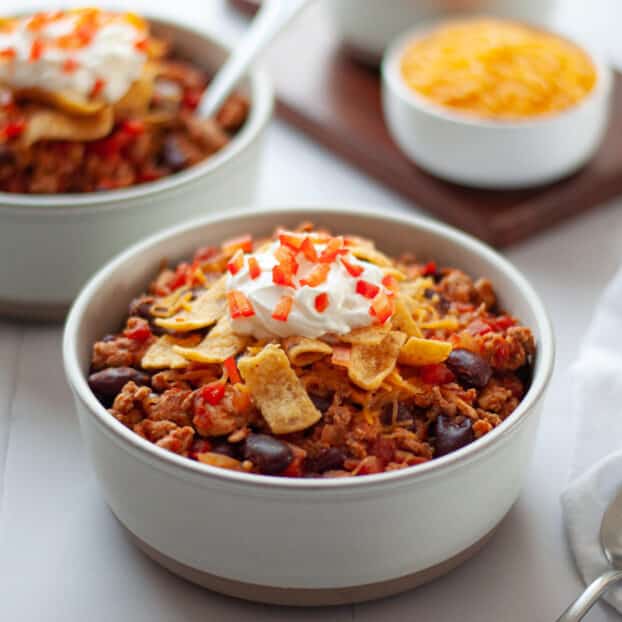 a stoneware bowl filled with healthy turkey chili topped with cheese, frito chips, sour cream, and diced red bell peppers. Another bowl of chili sits in the background, along with a small bowl of cheese and bowl of fritos on a wooden board. a white linen and spoons sit in frame too.