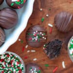an oreo truffle dipped in dark chocolate and topped with red, white, and green sprinkles next to another truffle with a bite out of it. The oreo ball is surrounded by additional truffles, sprinkles, bowls of sprinkles, and a white dish filled with oreo balls.
