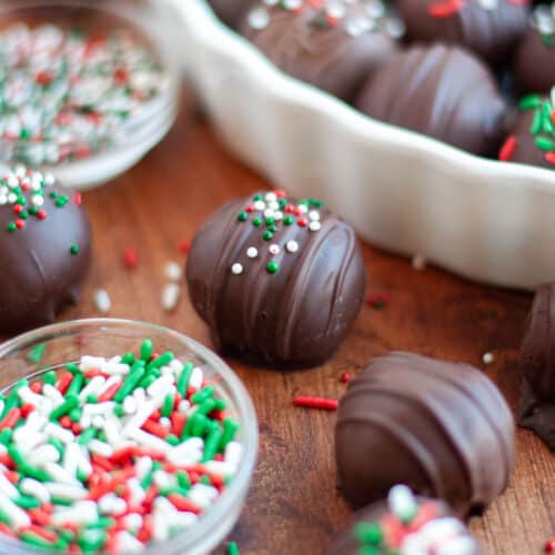 an oreo truffle dipped in dark chocolate and topped with red, white, and green sprinkles. The oreo ball is surrounded by additional truffles, sprinkles, bowls of sprinkles, and a white dish filled with oreo balls.