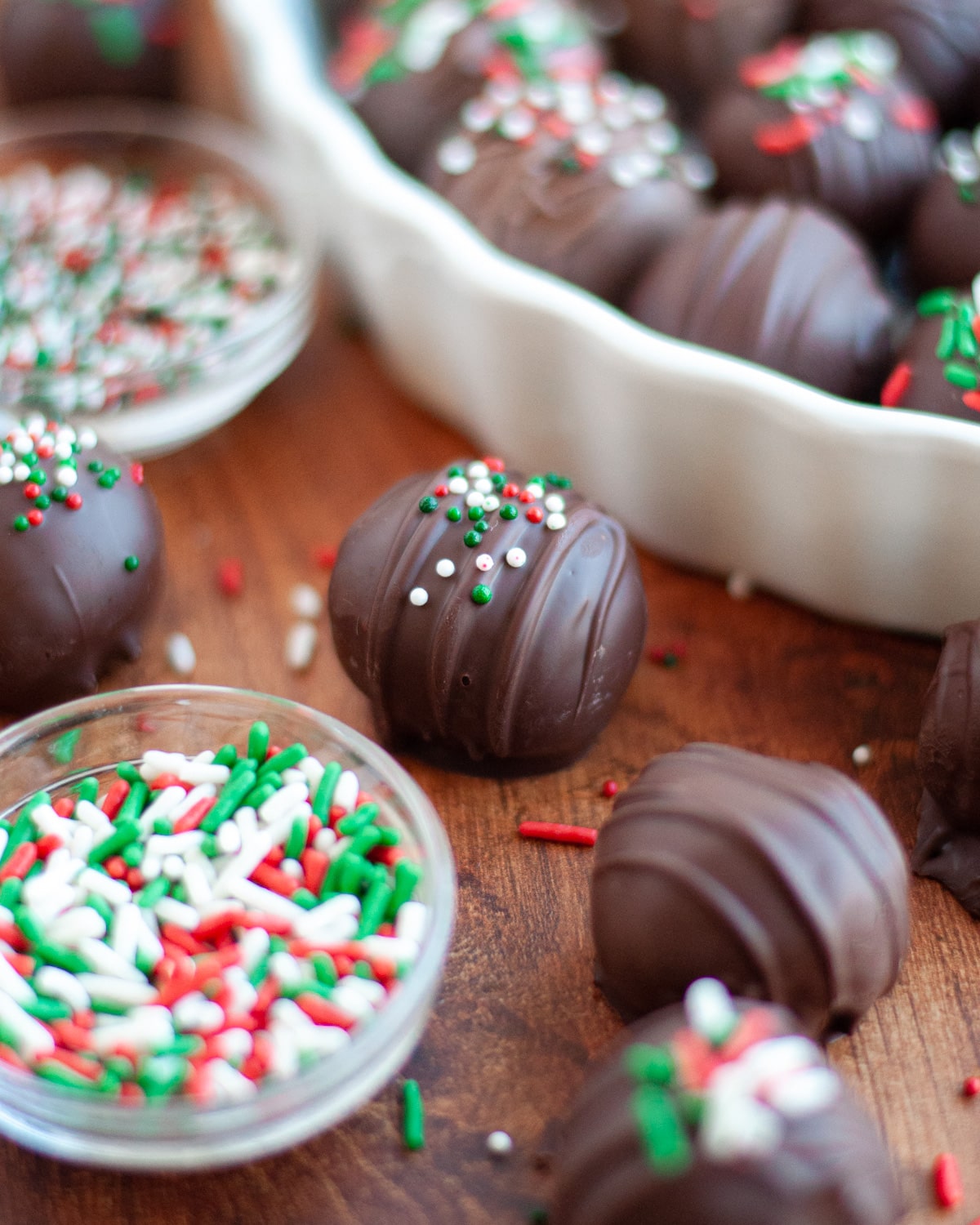 an oreo truffle dipped in dark chocolate and topped with red, white, and green sprinkles. The oreo ball is surrounded by additional truffles, sprinkles, bowls of sprinkles, and a white dish filled with oreo balls.