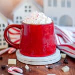 a peppermint white chocolate mocha in a red mug that is topped with whipped cream and crushed candy cane pieces. The mug sits on a white coaster in front of white holiday houses and a red and white linen. White chocolate pieces, coffee beans, and candy canes are scattered around the scene.