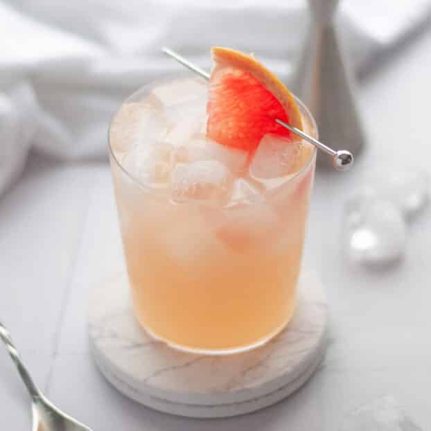 a pink grapefruit mocktail sitting on two white marble coasters with a slice of pink grapefruit garnishing the drink. there are ice cubes around the scene, as well as equipment used to make the drink like a jigger and bar spoon.