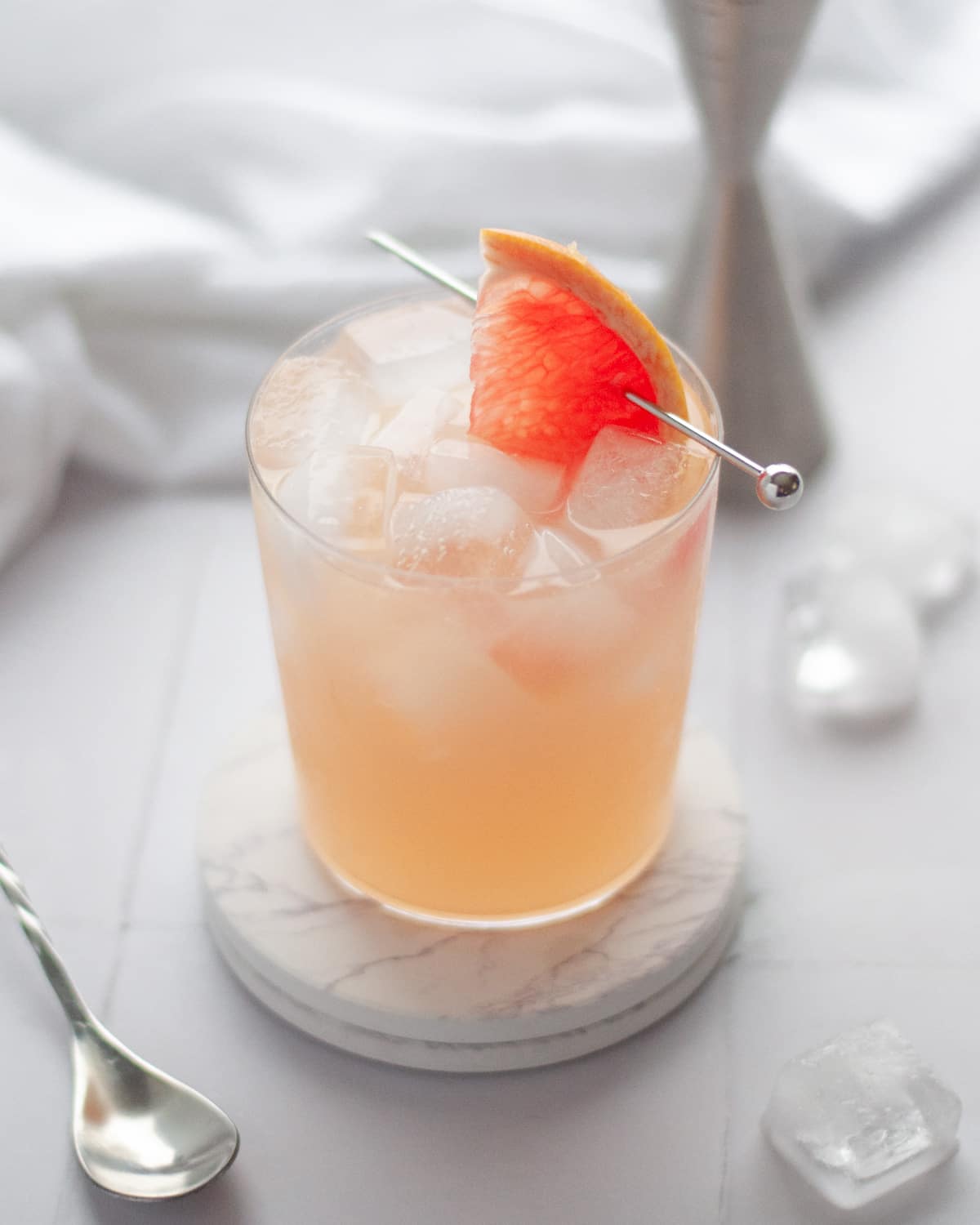a pink grapefruit mocktail sitting on two white marble coasters with a slice of pink grapefruit garnishing the drink. there are ice cubes around the scene, as well as equipment used to make the drink like a jigger and bar spoon.