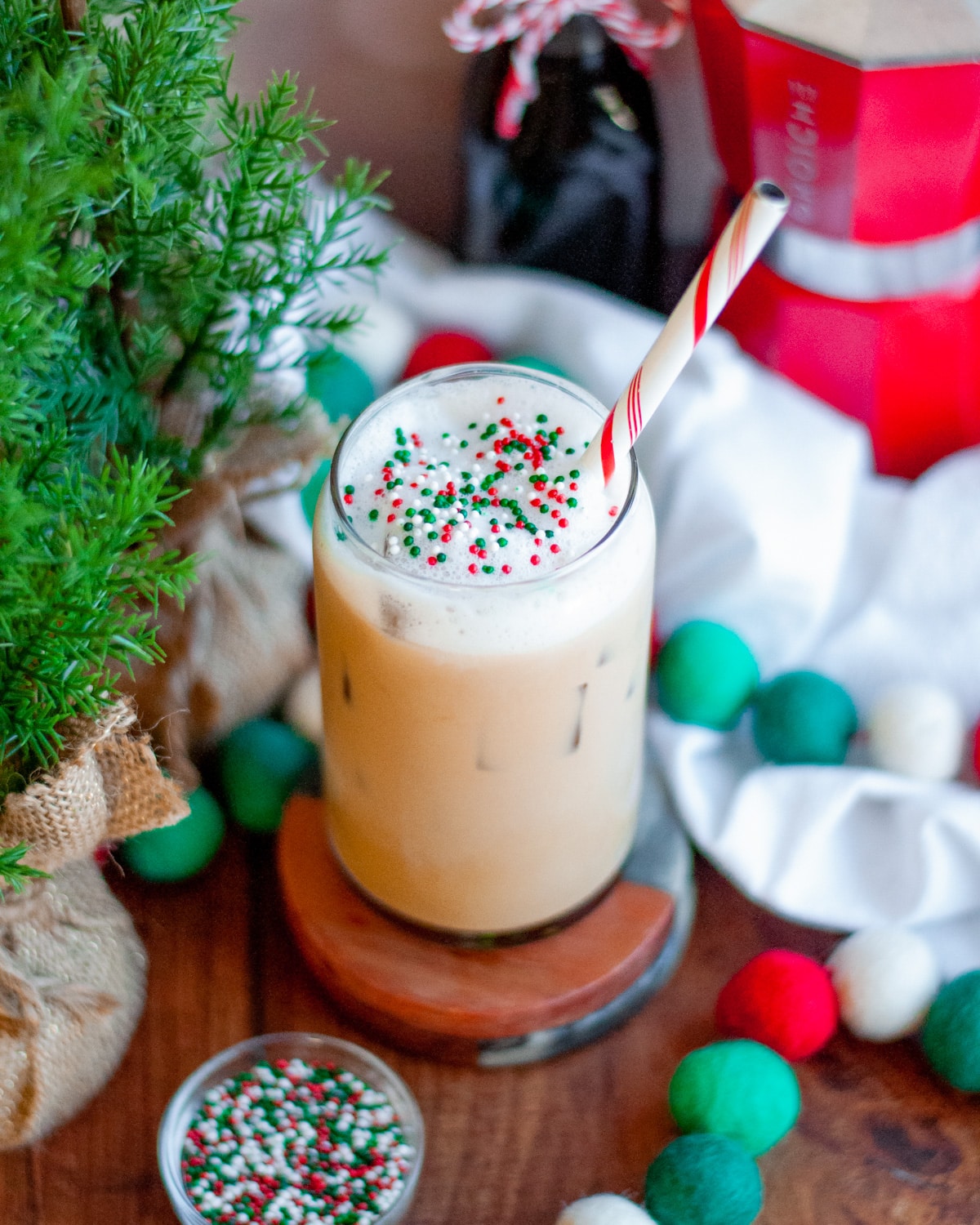 an iced sugar cookie latte with a red and white striped straw, and topped with red white and green sprinkles. The glass is surrounded by holiday felt-ball garland, containers of sprinkles, a jar of sugar cookie simple syrup with a bow, a red moka pot, a white linen, and greenery.