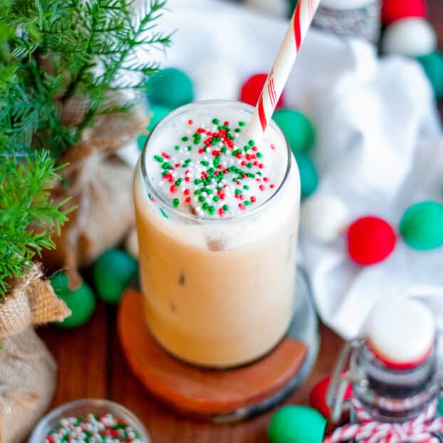 an iced sugar cookie latte with a red and white striped straw, and topped with red white and green sprinkles. The glass is surrounded by holiday felt-ball garland, containers of sprinkles, a jar of sugar cookie simple syrup with a bow, a white linen, and greenery.