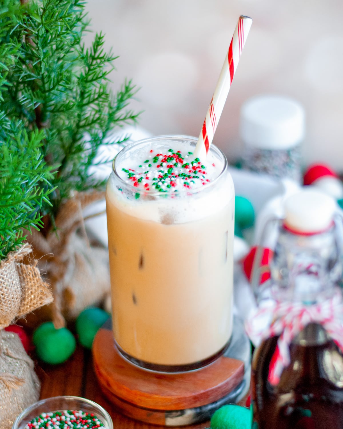 an iced sugar cookie latte with a red and white striped straw, and topped with red white and green sprinkles. The glass is surrounded by holiday felt-ball garland, containers of sprinkles, a jar of sugar cookie simple syrup with a bow, a white linen, and greenery.