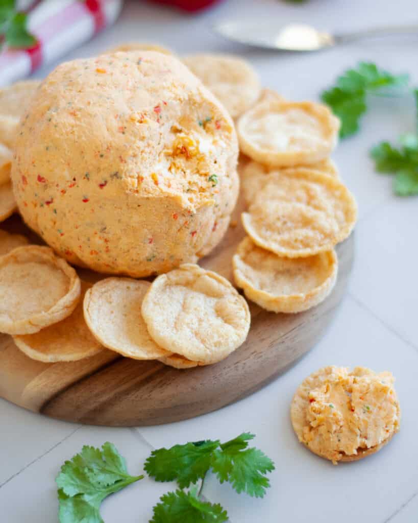 a tex mex cheese ball served on a round wooden platter with chips. there is a big scoop taken out of the cheeseball, and cilantro around the scene.