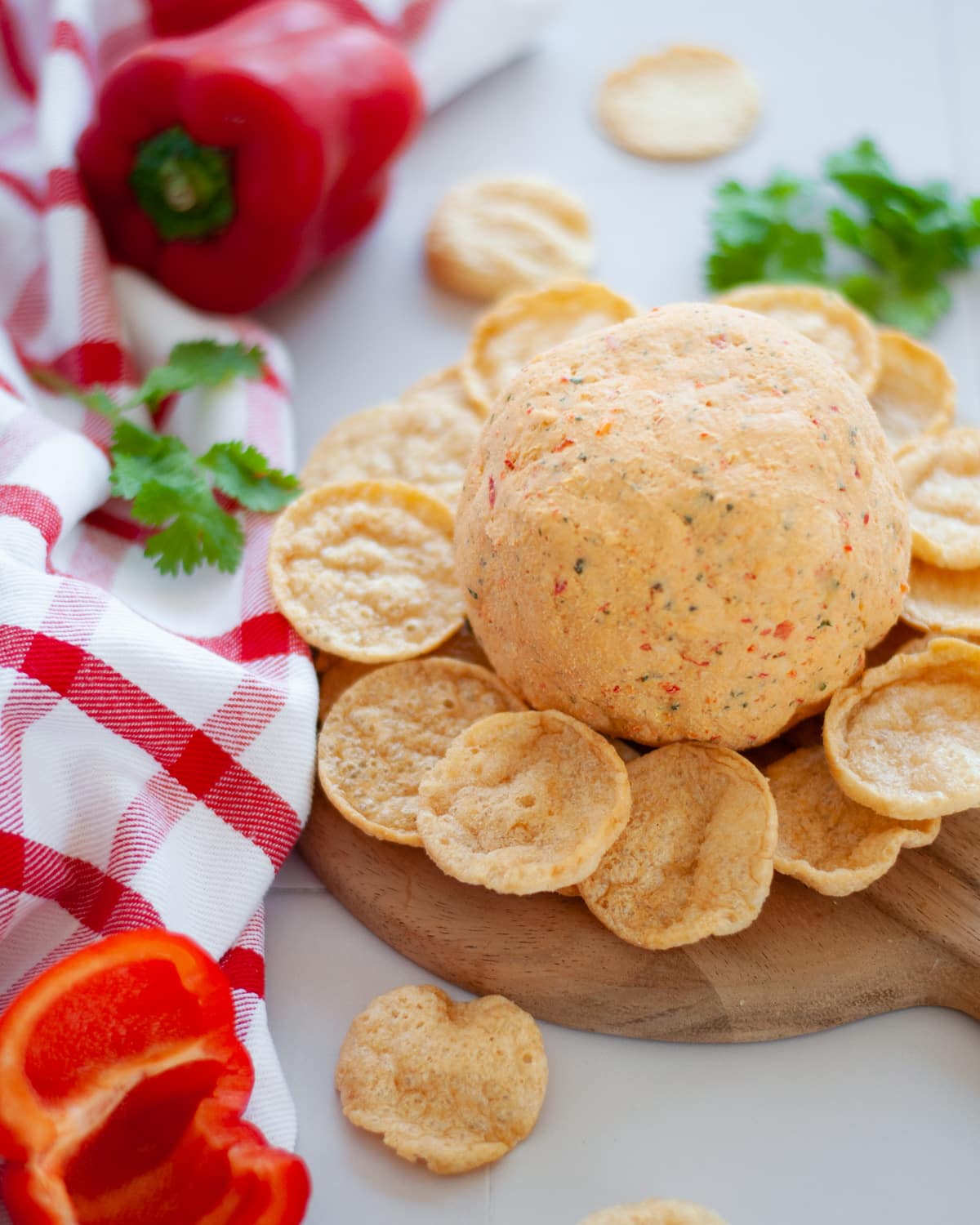 a tex mex cheese ball served on a round wooden platter with chips. there is cilantro, bell peppers, a red and white checked linen, and additional chips, around the scene.