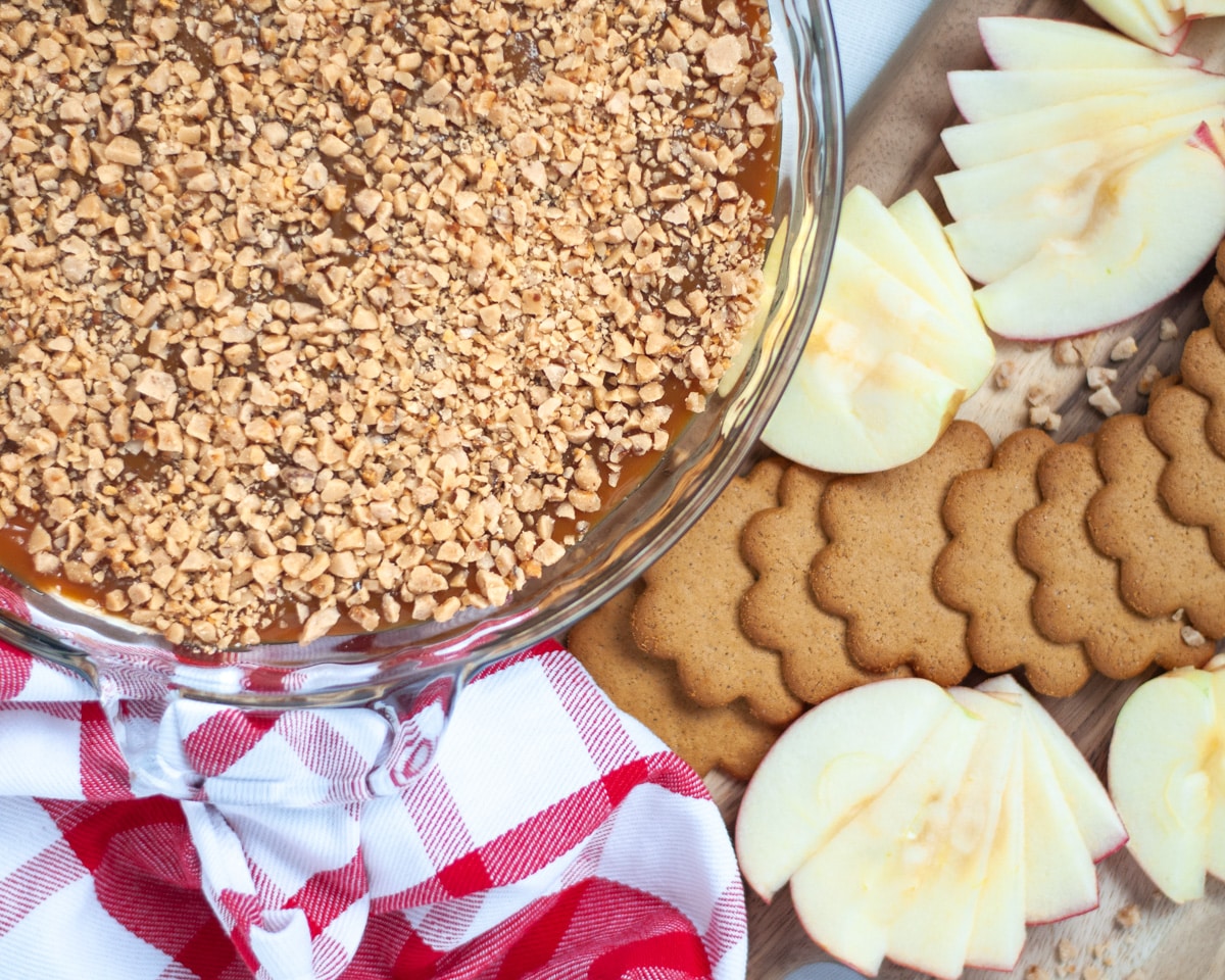 top down view of a glass pie pan filled with homemade caramel apple dip with toffee bits. Next to it is a board covered with gingersnap cookies and fresh apple slices as well as a red and white linen.