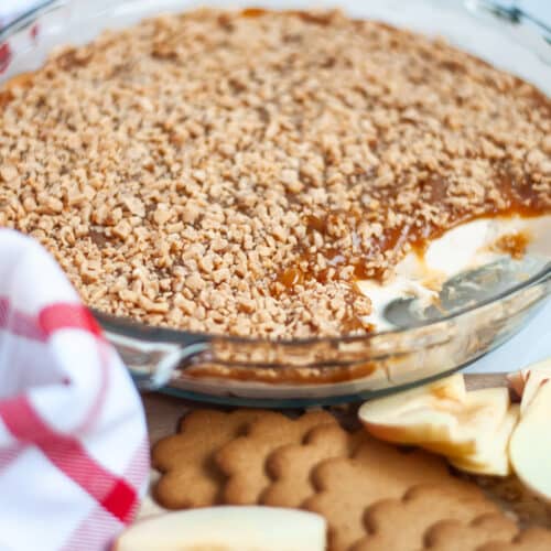 a deep glass pie pan filled with layered caramel apple dip with toffee bits with several scoops taken out of it. apple slices, gingersnaps, and a linen are in the forefront of the scene.