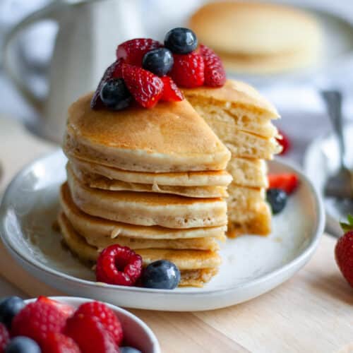 a big stack of fluffy oat milk pancakes topped with berries and maple syrup. the pancakes sit on a plate on a wood board with a bowl of berries in the foreground and a carafe of maple syrup and another plate of dairy free pancakes in the background.