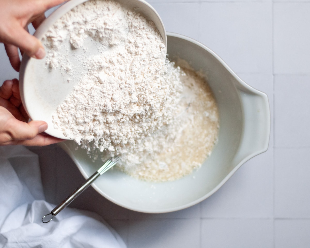 process shot showing how to make oat milk pancakes. this image shows the flour being added to the wet ingredients in a large mixing bowl.