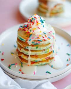 A big stack of funfetti pancakes with vanilla icing, whipped cream, and rainbow sprinkles on them. the stack of pancakes sits on a white plate, with another plate of pancakes in the background, and a white linen in front.