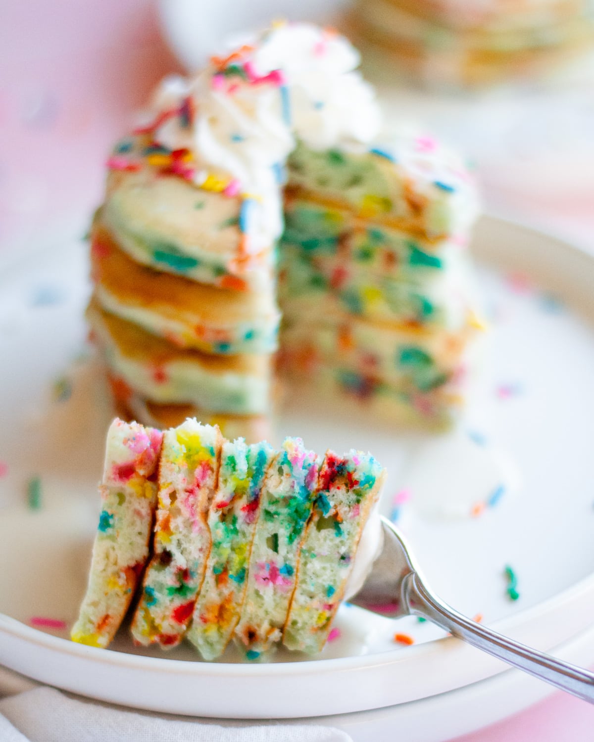 Close up of a fork full of spices of pancakes just removed from a big stack of funfetti pancakes with vanilla icing, whipped cream, and rainbow sprinkles on them. the stack of pancakes sits on a white plate, with another plate of pancakes in the background.