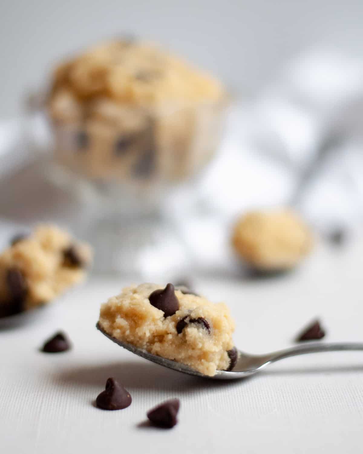 a spoonful of high protein edible cookie dough. the spoon of cookie dough is surrounded by chocolate chips and there are more spoons of cookie dough and a bowl of cookie dough blurred in the background.