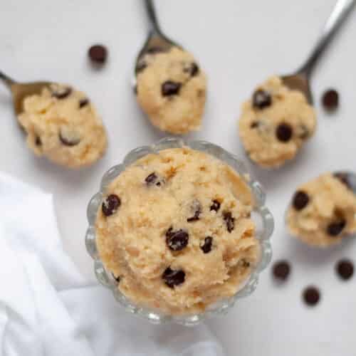 a glass bowl of high protein cookie dough surrounded by spoons full of edible cookie dough, chocolate chips, and a white linen.