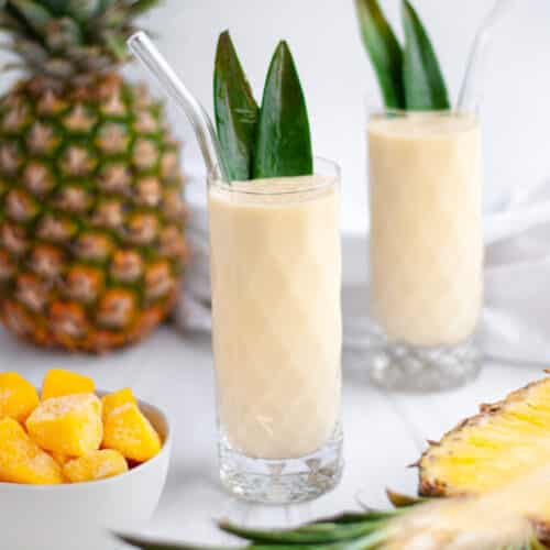 a tall serving glass filled with a fresh mango pineapple smoothie. the glass is garnished with 2 pineapple leaves and has a glass straw. the glass is surrounded by fresh pineapples, a bowl of mango pieces, a second smoothie, and a white linen.