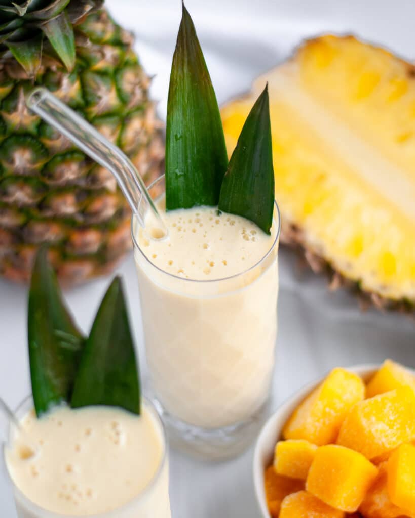 a tall serving glass filled with a fresh mango pineapple smoothie. the glass is garnished with 2 pineapple leaves and has a glass straw. the glass is surrounded by fresh pineapples, a bowl of mango pieces, a second smoothie, and a white linen.