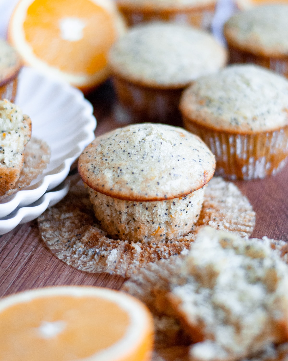 an unwrapped orange and poppy seed muffin sits on it's wrapper surrounded by more muffins, a stack of white plates, and cut oranges.