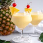 a fancy serving glass filled with a virgin pina colada mocktail with a pineapple wedge and maraschino cherry garnish. a fresh pineapple sits behind it, along with a second mocktail, a white linen, and tropical green leaves.