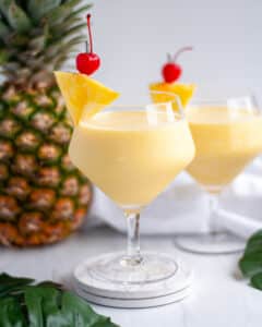 a fancy serving glass filled with a virgin pina colada mocktail with a pineapple wedge and maraschino cherry garnish. a fresh pineapple sits behind it, along with a second mocktail, a white linen, and tropical green leaves.