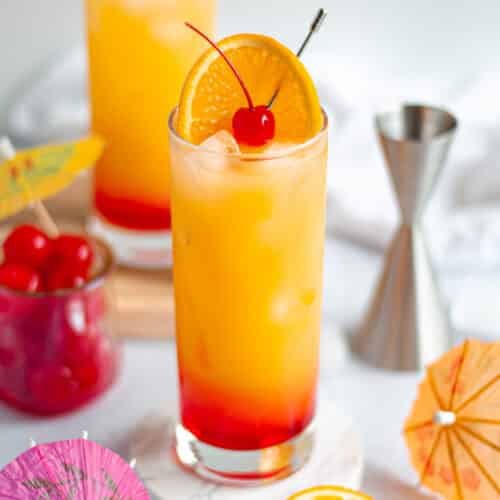 highball glass filled with a vodka sunrise cocktail. the glass is garnished with an orange slice and maraschino cherry on a cocktail pick. the glass is surrounded by additional fruit, drink umbrellas, a jigger, and another drink on a wooden board.