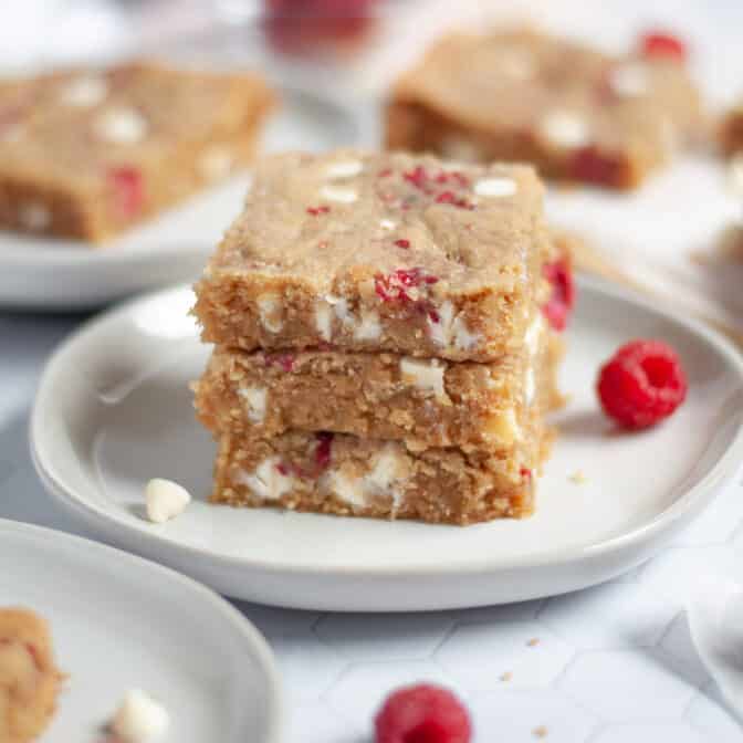 a stack of 3 white chocolate raspberry blondie squares on a plate. additional blondies surround the stack, and white chocolate chips and fresh raspberries are scattered around the scene.