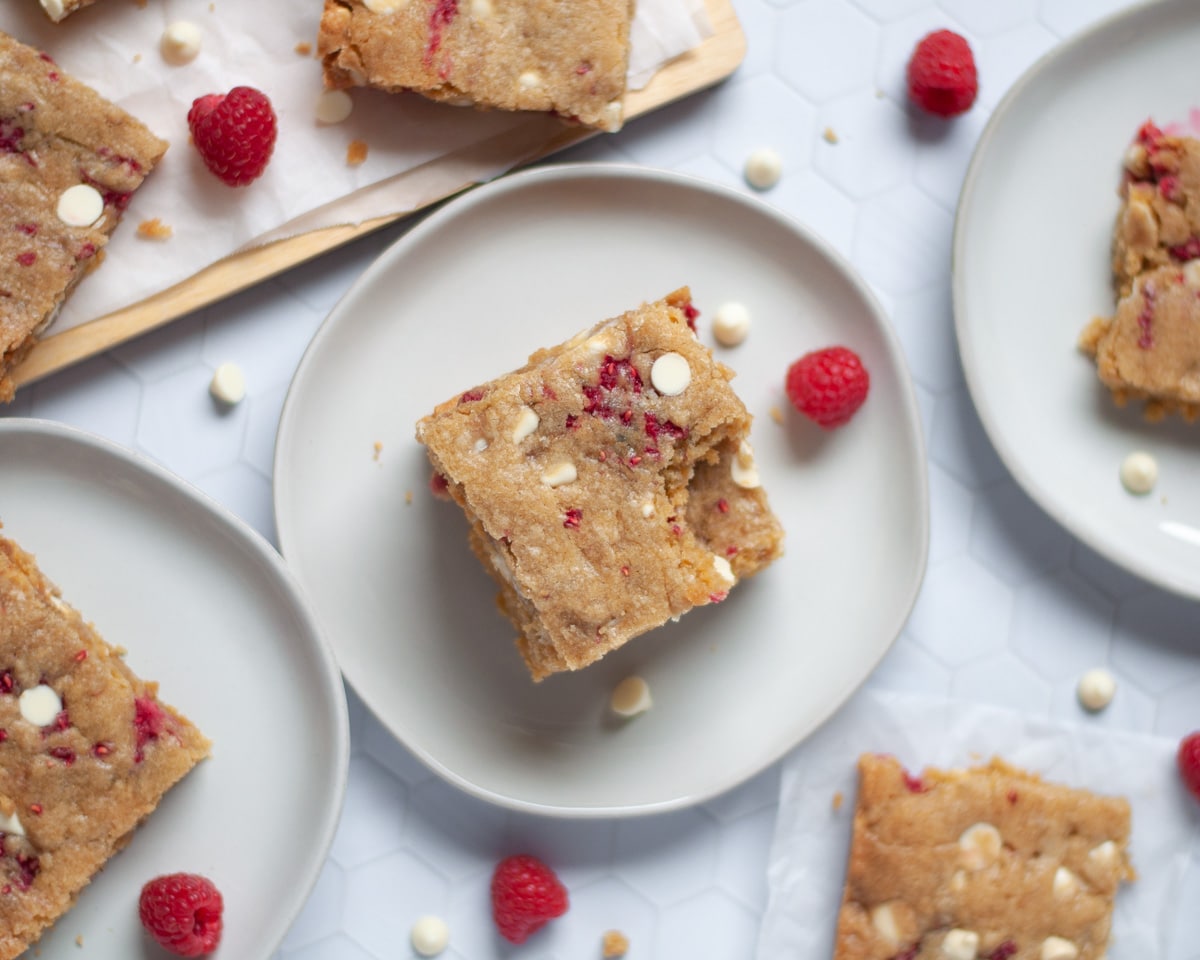 a stack of raspberry white chocolate blondie squares on a plate, with the top bar having a bite out of it. additional blondies surround the stack, and white chocolate chips and fresh raspberries are scattered around the scene.
