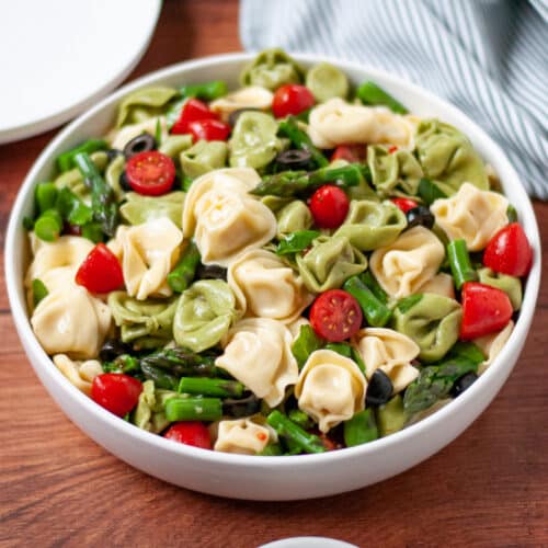 a large serving bowl filled high with summer tortellini salad with Italian dressing. A white serving spoon, white plates, and a green and white linen sit around the scene.