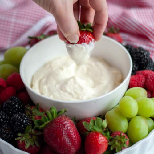 a hand dunking a strawberry into a bowl of 2-ingredient fruit dip that sits on a fruit tray with a variety of fresh fruit.