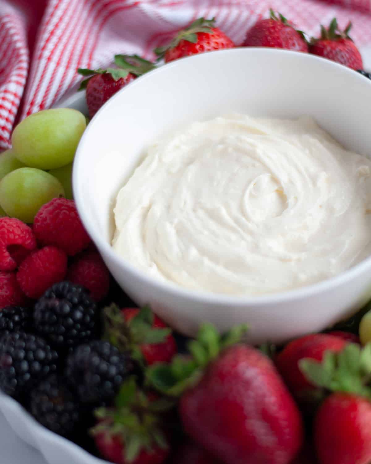 2 ingredient fruit dip in a white bowl, in the middle of a tray full of fresh fruit. The tray has green grapes, strawberries, blackberries, and raspberries.