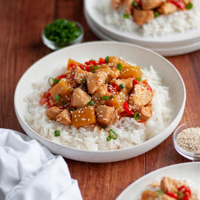 serving of sweet hawaiian chicken in crockpot served over rice in a white dish. the chicken is topped with green onions and green onions and sits next to additional plates of the dish served and ready to eat, toppings in small bowls, and a white linen.