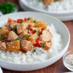 close up of sweet hawaiian crockpot chicken served over rice in a white dish. the chicken is topped with green onions and green onions. additional plates are in the foreground and background, and small dishes of toppings too.