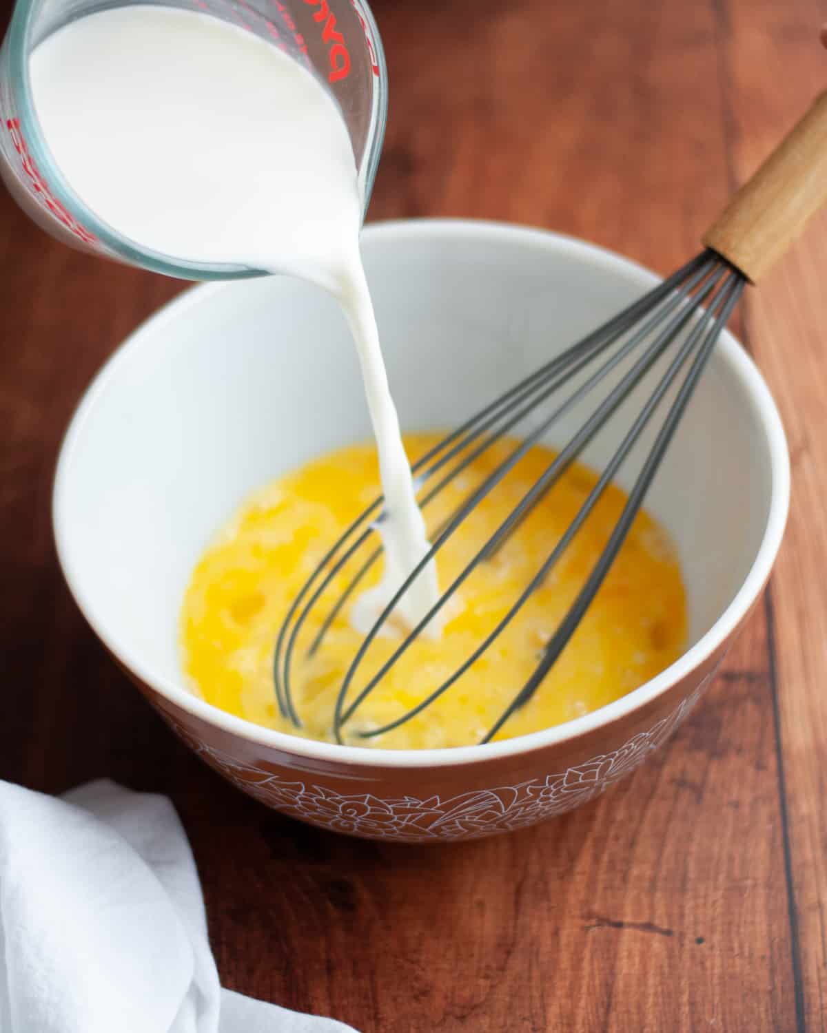 process shot showing how to make the custard for brioche french toast bake. image shows milk and cream being poured into whisked eggs in a medium mixing bowl with a whisk.
