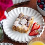 a slice of brioche french toast casserole sitting on a beautiful serving plate with sliced strawberries. the serving dish of the french toast bake sits in the back, with a bowl of berries, glasses of juice, and a vase of flowers around the scene.