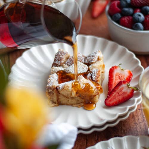 maple syrup being poured onto a slice of brioche french toast casserole sitting on a beautiful serving plate with sliced strawberries. the serving dish of the french toast bake sits in the back, with a bowl of berries, glasses of juice, and a vase of flowers around the scene.