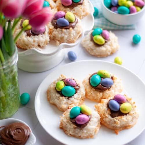 a white plate + a white cake stand filled with coconut macaroon nests. Additional easter coconut macaroons, and small bowls of nutella and m&m eggs. pink tulips and a blue and white checkered linen are also in the scene.