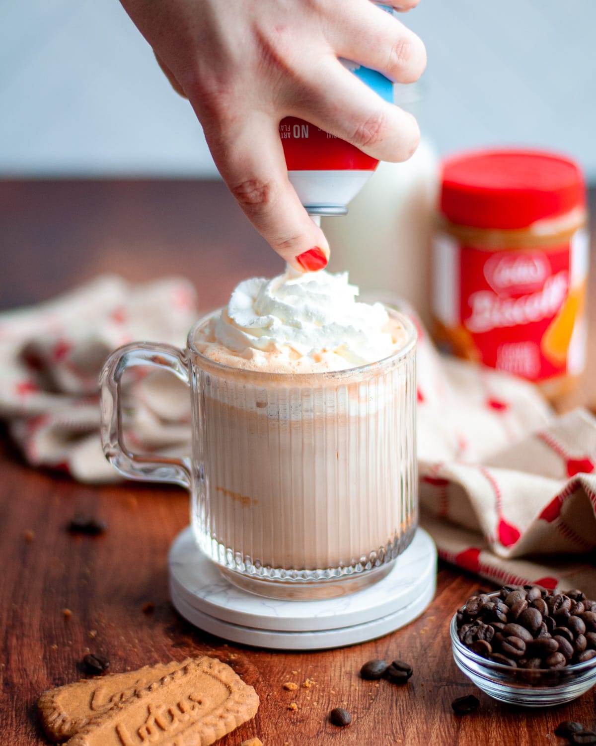 process shot showing whipped cream being added on top of the cookie butter latte. the mug sits on top of 2 white coasters and is surrounded by a tan and red linen, lotus biscoff cookies, and coffee beans. a container of milk and the jar of cookie butter sit in the background.