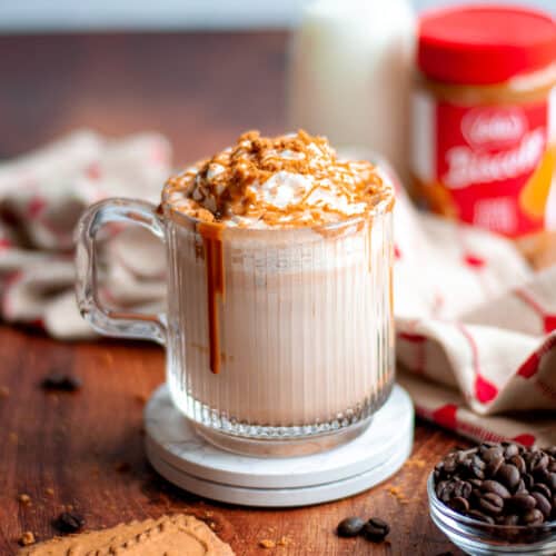 a delicious cookie butter latte in a glass mug. the mug sits on top of 2 white coasters and is surrounded by a tan and red linen, lotus biscoff cookies, and coffee beans. a container of milk and the jar of cookie butter sit in the background.