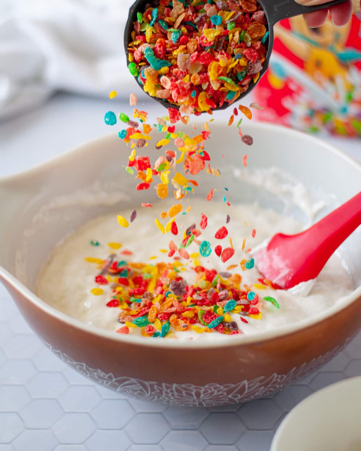 fruity pebbles being added to a large mixing bowl filled with melted mini marshmallows. a box of fruity pebbles cereal and a white linen sit behind it.