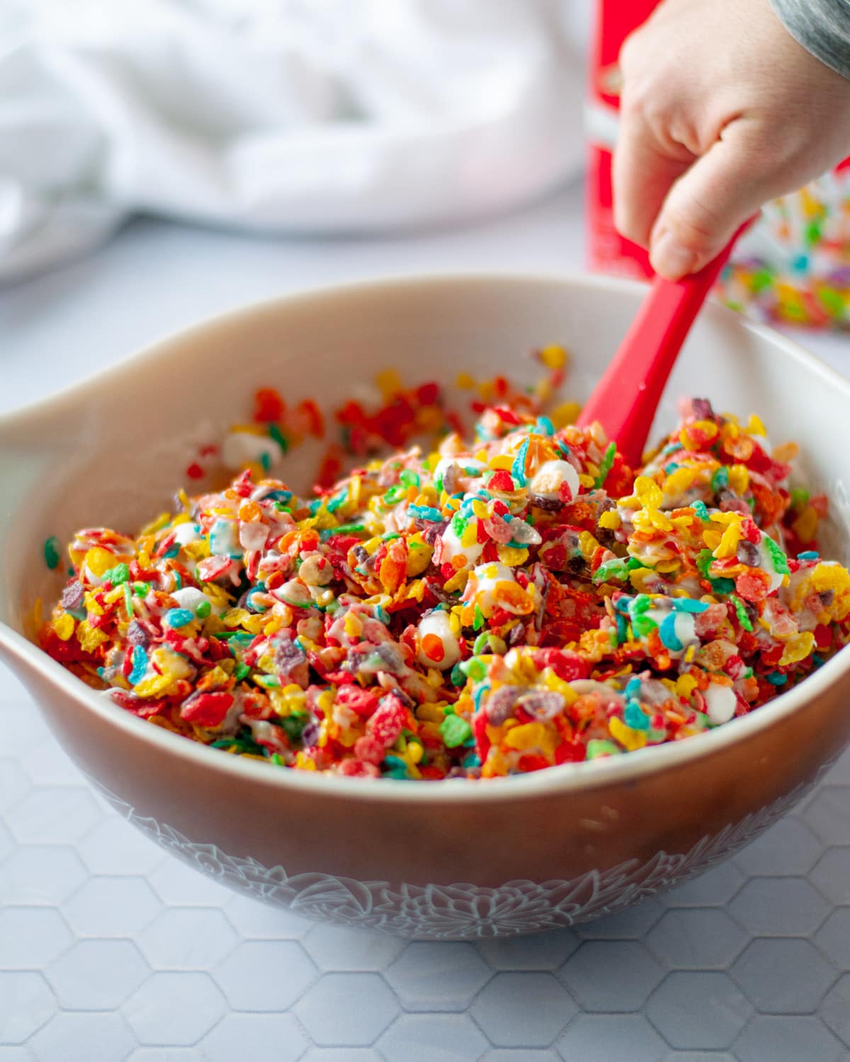 fruity pebbles and mini marshmallows being mixed with melted marshmallows and butter in a large mixing bowl. a box of fruity pebbles cereal and a white linen sit behind it.