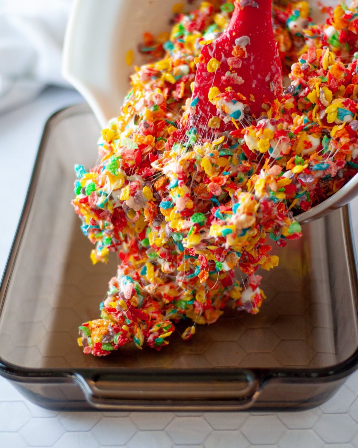 the fruity pebbles treats with marshmallows mixture being transferred from a large mixing bowl to a 9x13 pan.