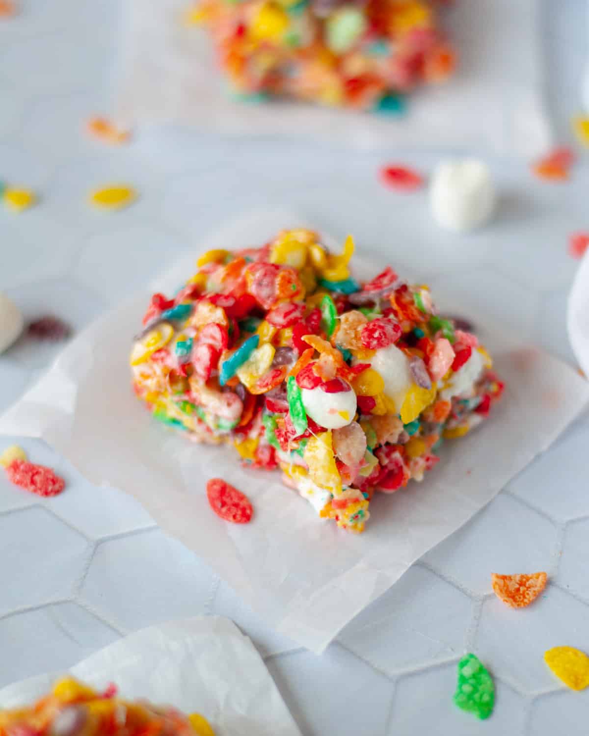 close up of a square of fruity pebbles marshmallow treats on a piece of parchment paper. fruity pebbles, mini marshmallows, and additional fruity pebbles bars.