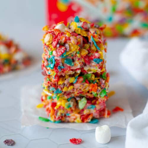 a stack of fruity pebbles cereal bars on a piece of parchment paper. fruity pebbles, mini marshmallows, and additional fruity pebbles bars. a box of fruity pebbles and a white linen sit in the background.