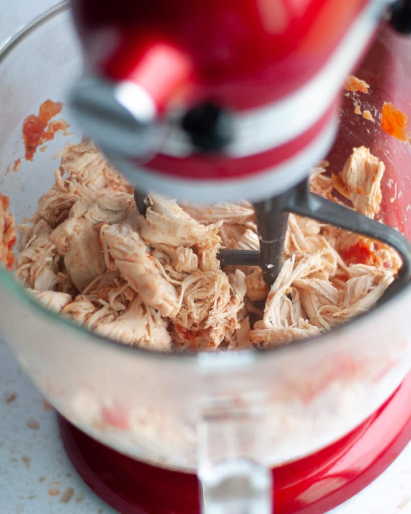 process shots showing how to make 2-ingredient crockpot salsa chicken. this image shows the cooked chicken being shredded with the paddle attachment in the bowl of a stand mixer.