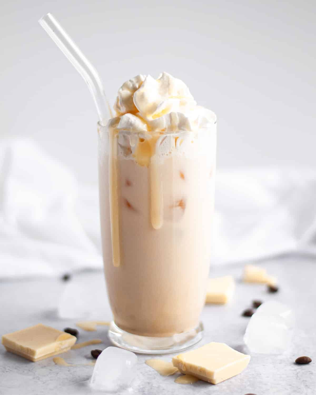 a white chocolate iced mocha in a tall glass, topped with shipped cream and white chocolate sauce with a few big drips of chocolate running down the glass. the glass is surrounded by ice cubes, white chocolate pieces, and coffee beans.