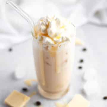 a white chocolate iced mocha in a tall glass, topped with shipped cream and white chocolate sauce with a few big drips of chocolate running down the glass. the glass is surrounded by ice cubes, white chocolate pieces, and coffee beans.
