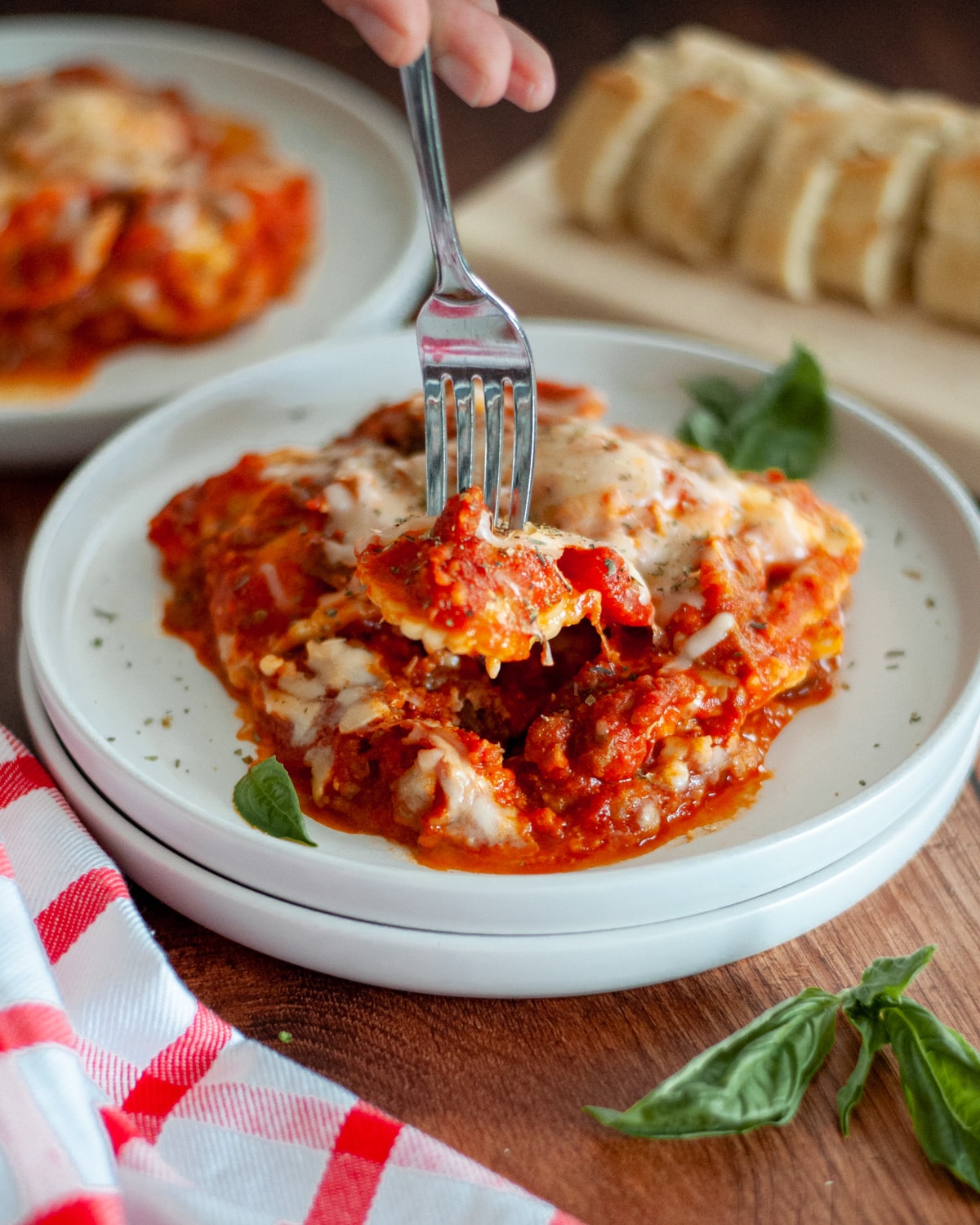 a white plate with a serving of crockpot lazy lasagna. a fork is digging into the serving and holds a ravioli. It is topped with Italian seasoning, and fresh basil leaves sit around as garnish.